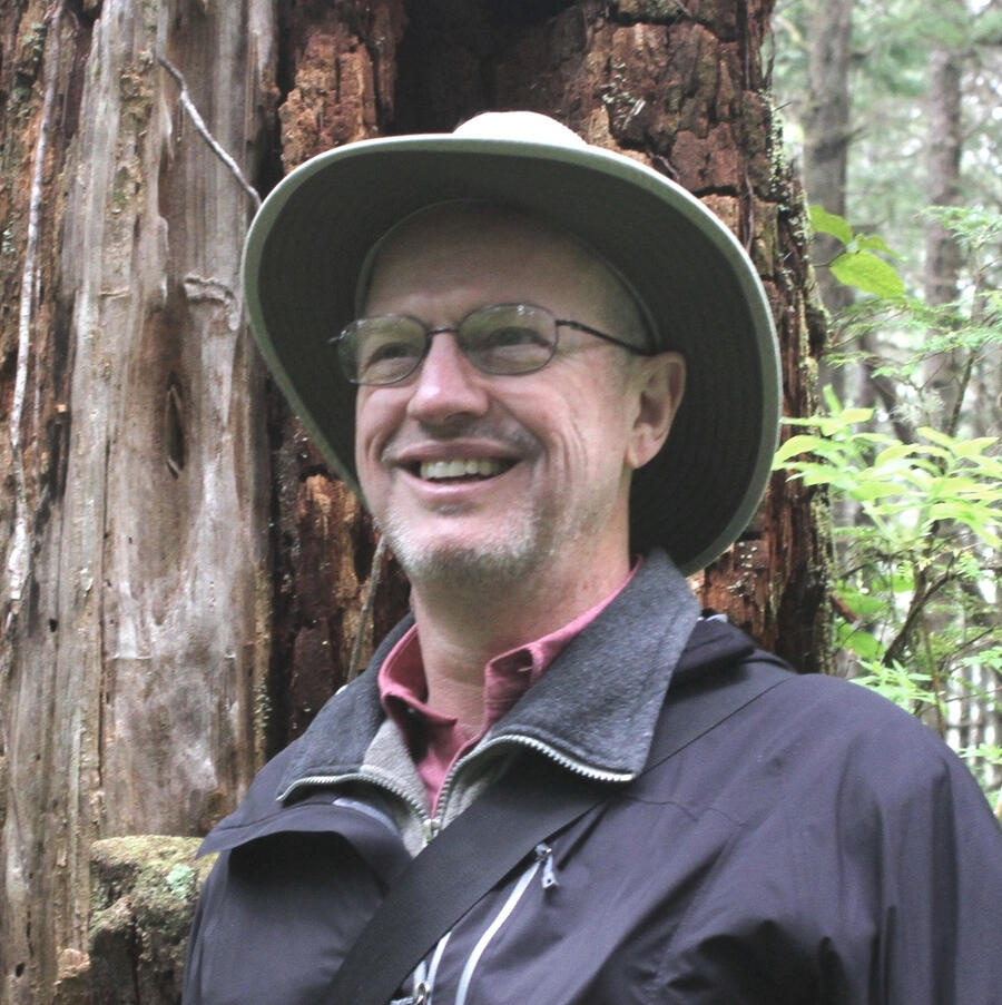 Thomas S. "Tom" Bremer portrait A close-up portrait of a smiling man with glasses and a wide-brimmed hat standing in front of a tree trunk.