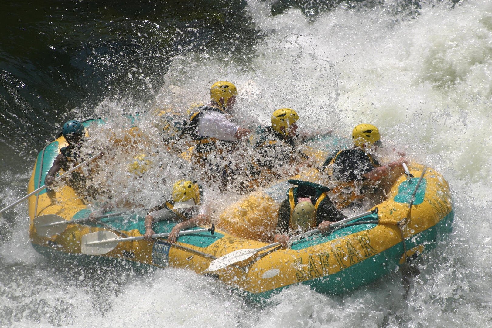 A group of people wearing yellow helmets and life jackets is white water rafting down the Elk River in a yellow inflatable boat, surrounded by splashing water as they navigate rapids.