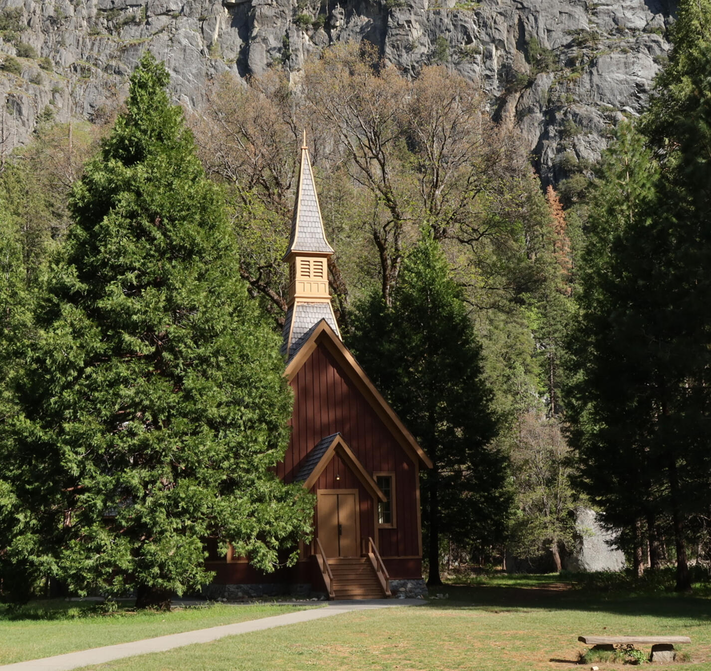 Yosemite Valley Chapel A small, brown wooden church with a tall steeple sits among large evergreen trees at the base of a rocky Yosemite cliff, with sunlight illuminating the building and patches of grass in the foreground.